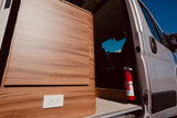 RV rental interior: sleek wood paneling with an outlet & essential fire extinguisher by the open door. Blue sky outside.