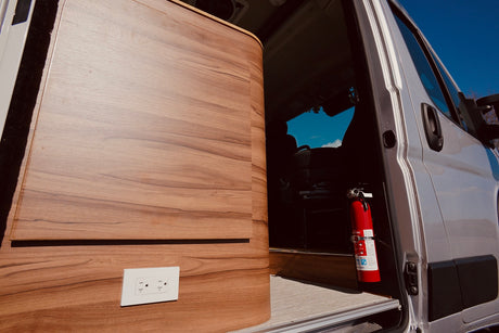 RV rental interior: sleek wood paneling with an outlet & essential fire extinguisher by the open door. Blue sky outside.