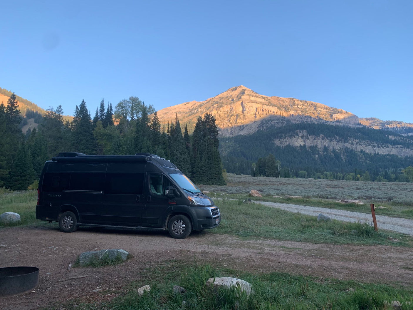 Black RV rental parked at a serene mountain campsite, illuminated by the warm glow of sunrise. Ideal for nature lovers.