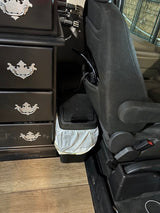 Inside an RV rental: a black dresser with silver pulls, passenger seat, and a small trash can with liner on wood flooring.