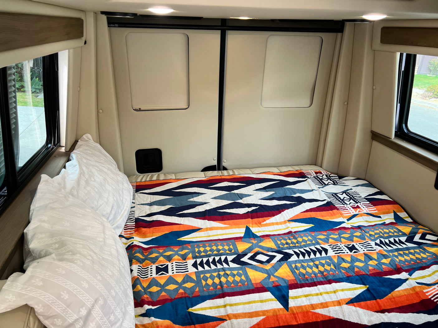 Cozy RV rental bedroom with a colorful geometric blanket, white pillows, and natural light from side windows.