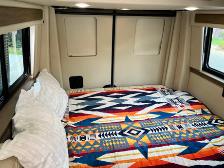 Cozy RV rental bedroom with a colorful geometric blanket, white pillows, and natural light from side windows.