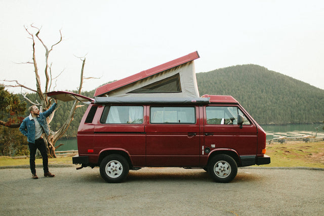 Red pop-top camper van, an ideal rv rental, parked by a scenic mountain lake. A man stands at the open rear hatch.