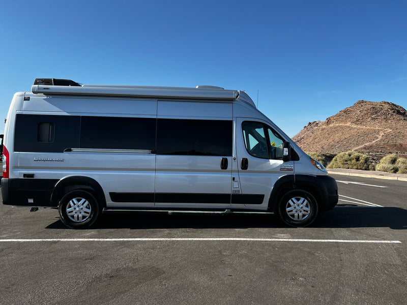 This silver Class B RV rental, a Ram ProMaster camper van, is parked on asphalt with a mountain in the distance under a blue sky. — photo 17