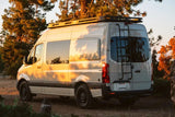 Light gray custom Sprinter van RV rental with roof rack & ladder, bathed in golden light in a forest setting.