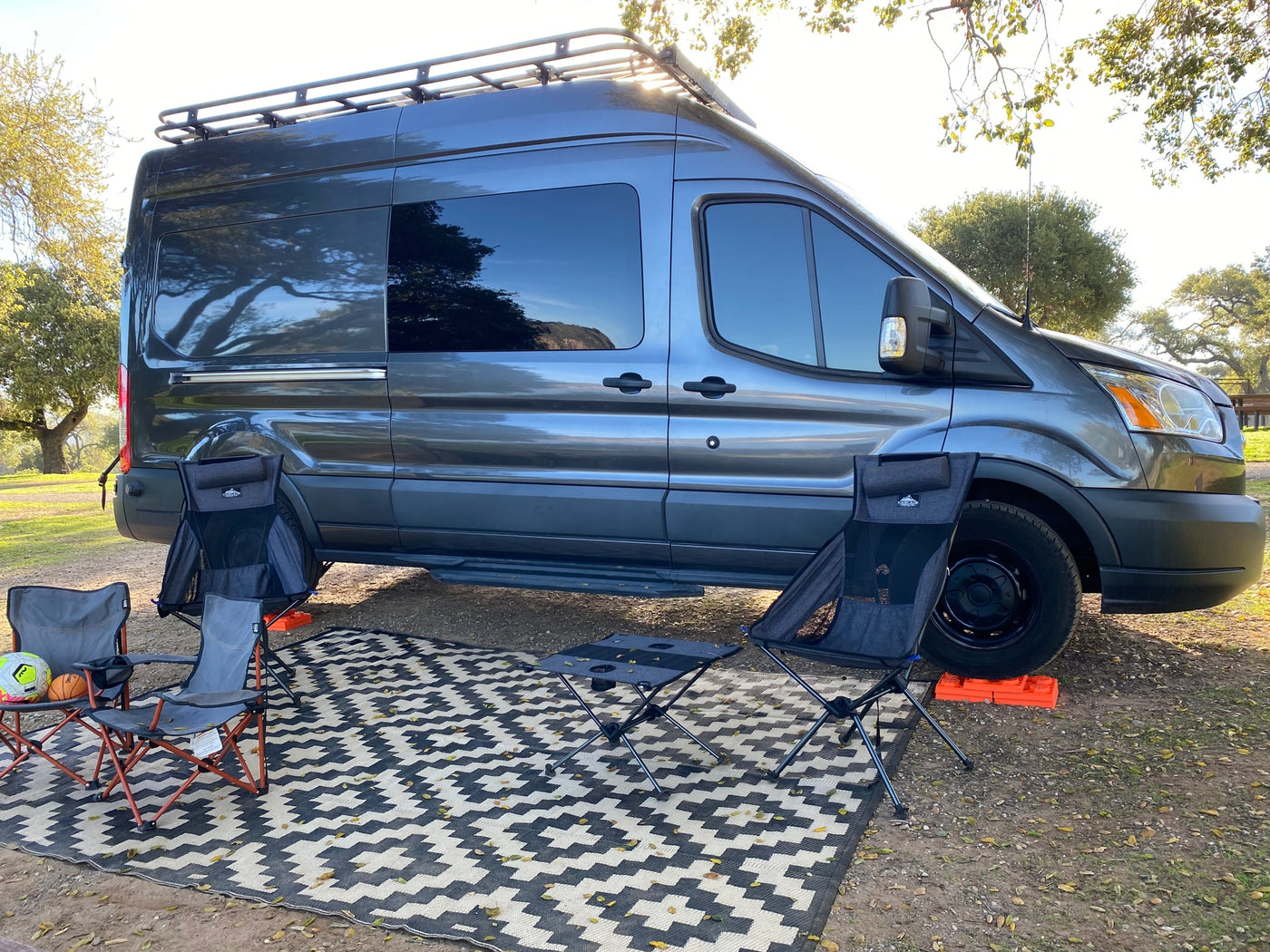 A dark gray camper van with a roof rack and leveling blocks, set up for camping with outdoor chairs and mats, ideal for rv rental adventures. — photo 25