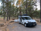White Ram ProMaster City rv rental camper van at a wooded campsite. Equipped with roof box & cooler for outdoor camping.