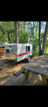 Small white RV rental camper with AC unit and red stripe by a picnic table, green cord visible. Camping by a lake.