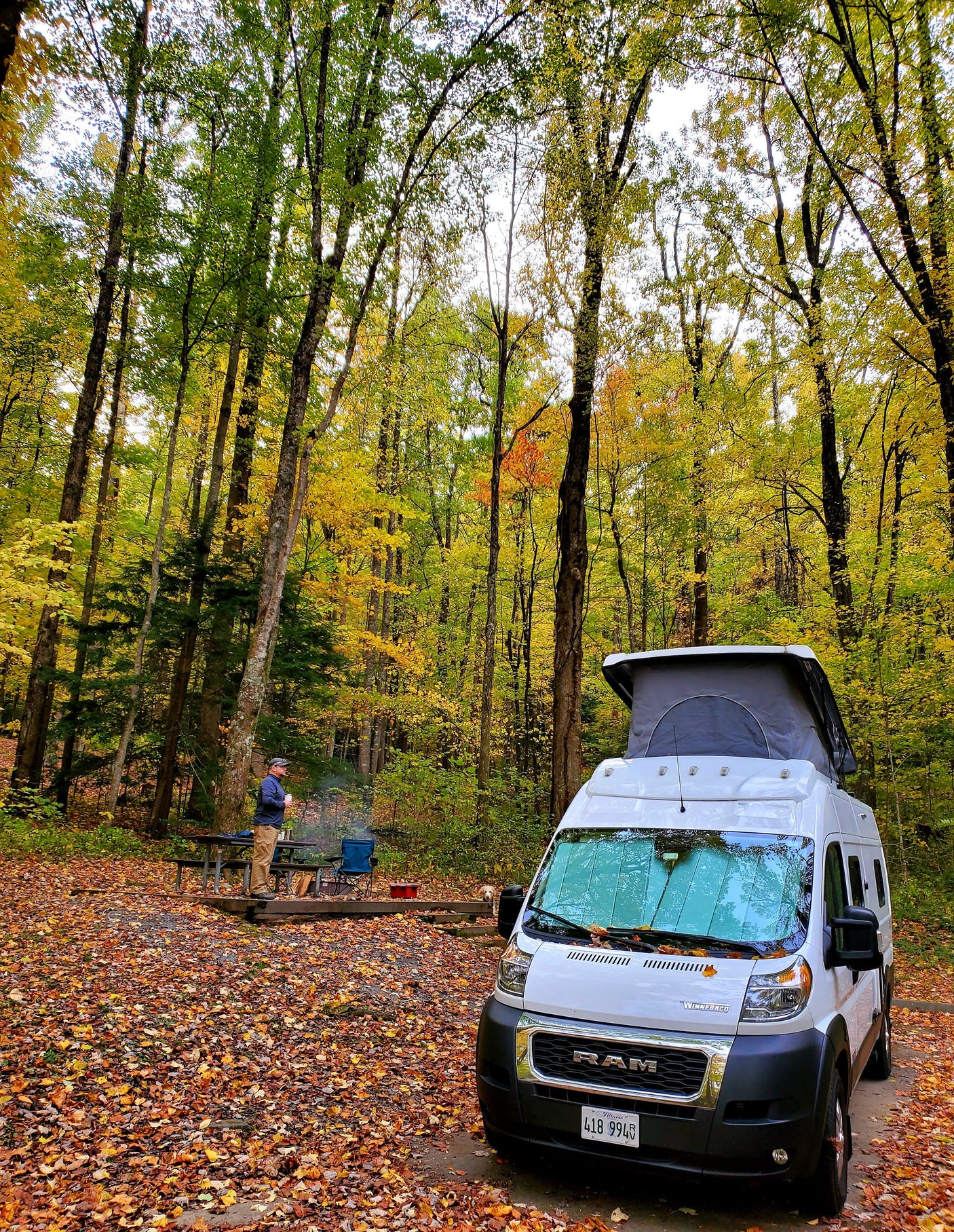 Enjoying autumn camping with an RV rental: a white camper van at a vibrant fall campsite, featuring a cozy campfire, person, and dog.