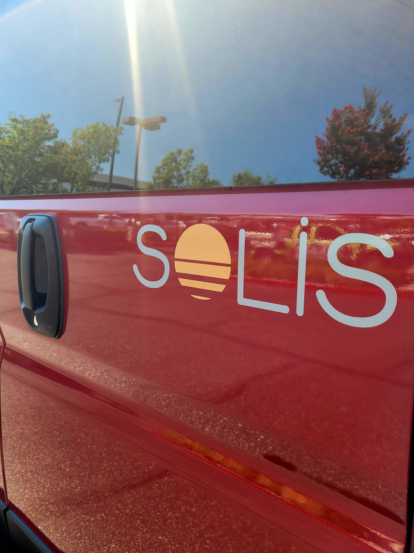 Red Solis RV rental side with sun-like logo, door handle, and blue sky reflection in window.