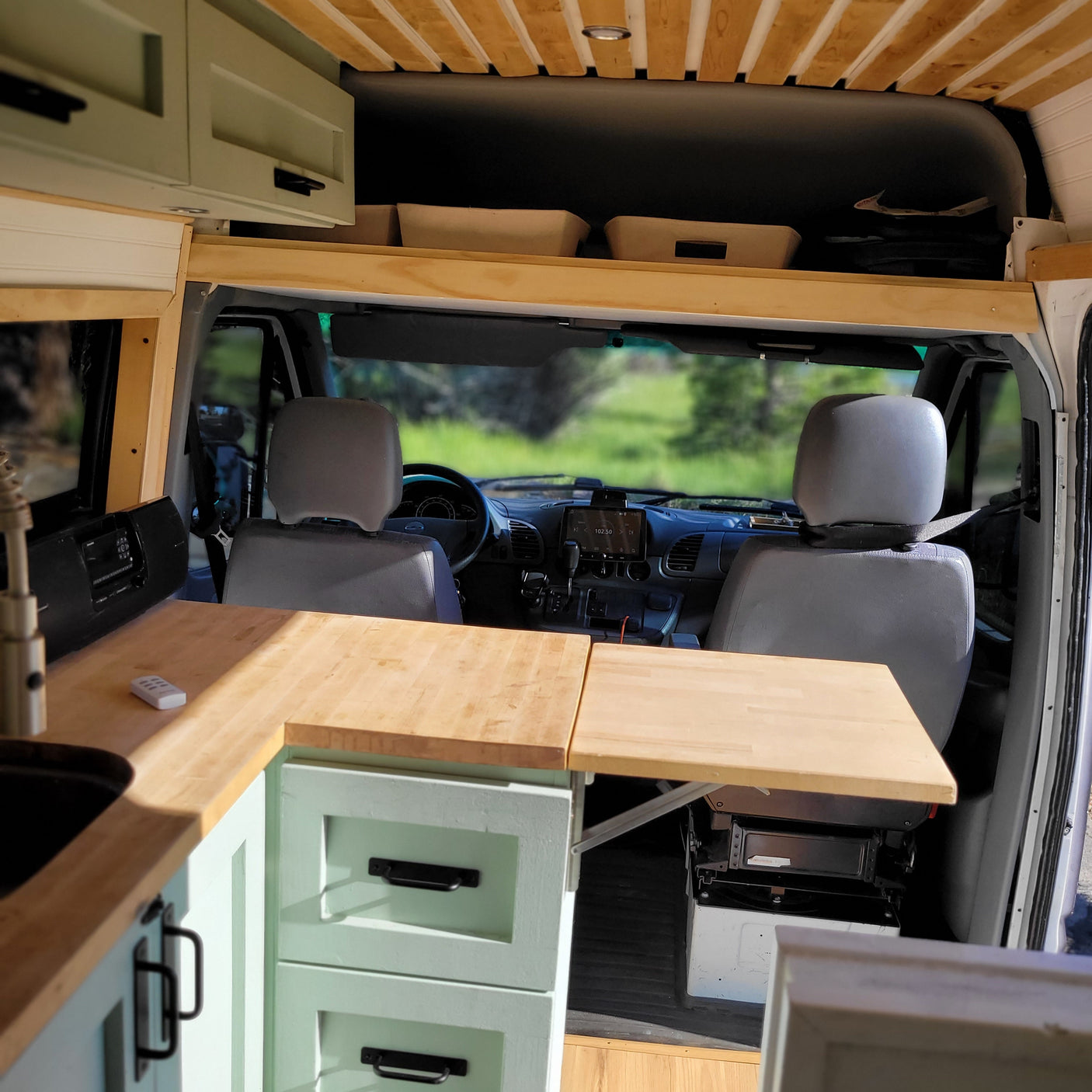 Inside this RV rental: a custom camper van with light wood ceiling, green cabinets, kitchen counter, & front cabin view.