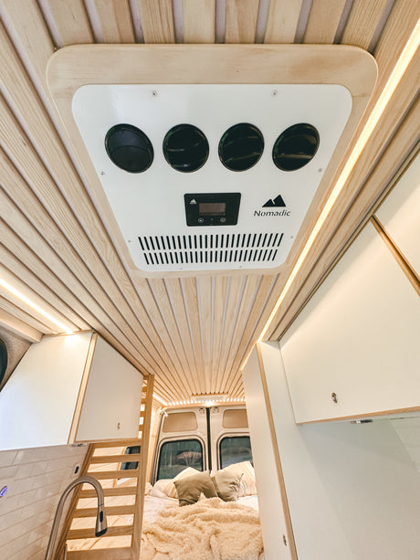 Bright RV rental interior: slatted wooden ceiling, Nomadic AC, ambient LED lighting, bed, and kitchen sink visible.