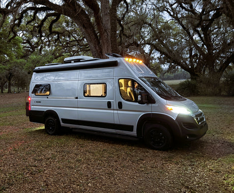 A silver campervan with lights on, parked under large, mossy trees at dusk. Ideal for your next rv rental adventure.