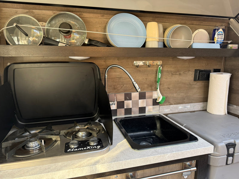 RV rental: Compact kitchen with stovetop, sink, dishes, pots, and a cooler, organized for cooking on the go. — photo 4