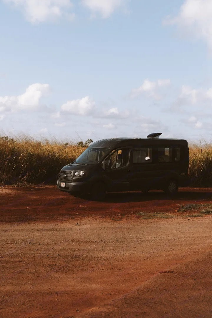 A dark Ford Transit camper van, an RV rental, parked on a red dirt road with dry grass under a blue sky.