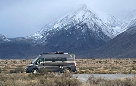 Gray RV rental parked on a vast plain with dramatic snow-capped mountains. Ideal for scenic road trips and wilderness adventures.
