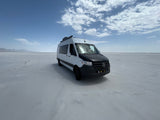 A white and black RV rental van parked on a vast salt flat, with distant mountains and a blue sky overhead.