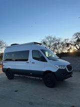 A white "Tranquility" Mercedes Sprinter camper van, an ideal rv rental, parked on a paved lot at sunset.