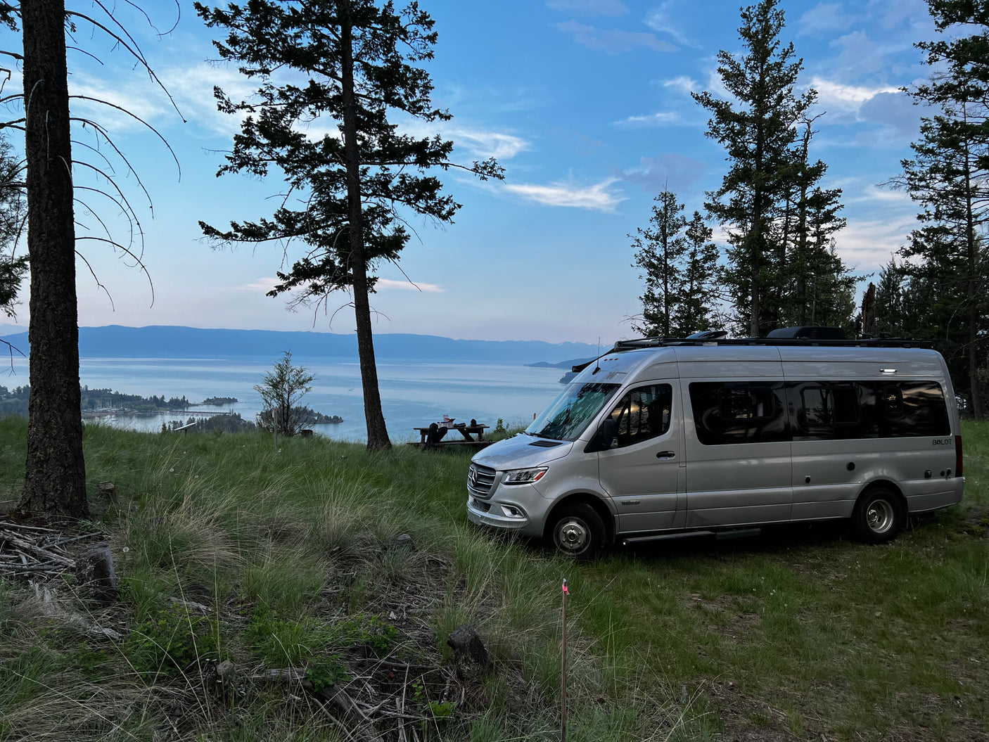 An RV rental, a silver Winnebago Boldt campervan, parked overlooking a scenic lake and mountains at dusk.