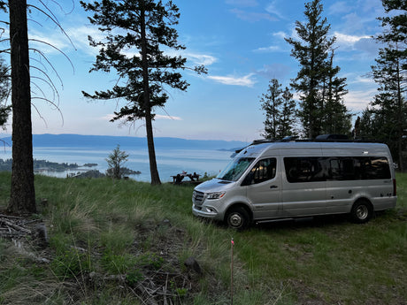 An RV rental, a silver Winnebago Boldt campervan, parked overlooking a scenic lake and mountains at dusk.