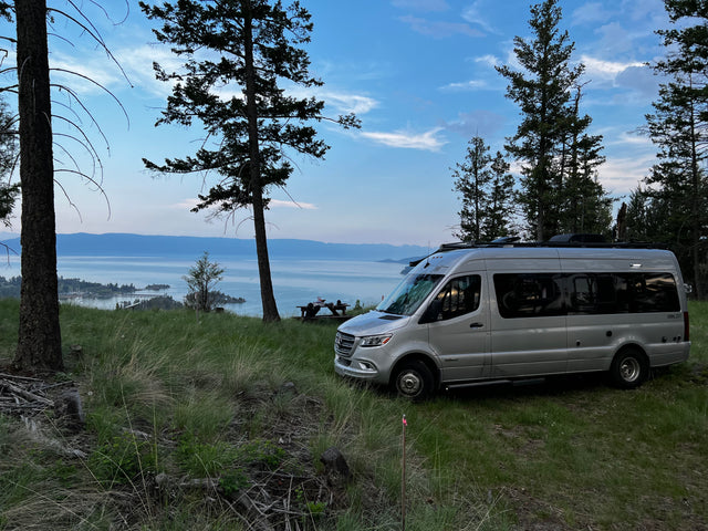 An RV rental, a silver Winnebago Boldt campervan, parked overlooking a scenic lake and mountains at dusk.