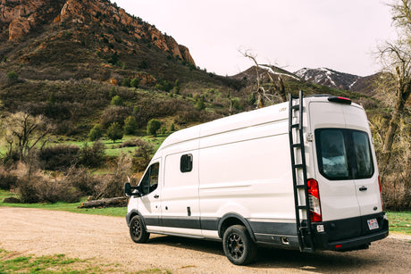 White rv rental camper van with a ladder, parked on a dirt road in a scenic mountainous landscape.