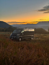 A dark rv rental camper van sits in tall, golden grass at sunset, with mountains framing the vibrant sky.