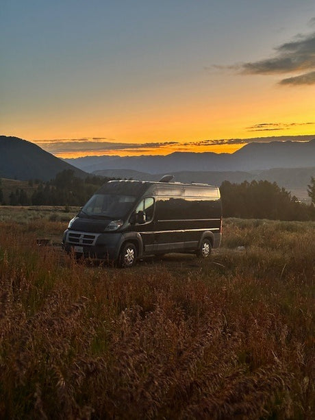 A dark rv rental camper van sits in tall, golden grass at sunset, with mountains framing the vibrant sky.