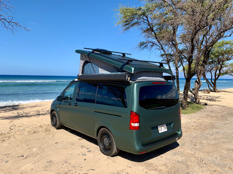 Dark green pop-top camper van RV rental parked on a sandy Hawaiian beach near the ocean.