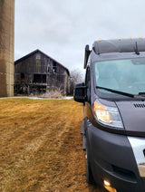 Grey RV rental van on dry grass, with a rustic, dilapidated barn and silo under an overcast sky.