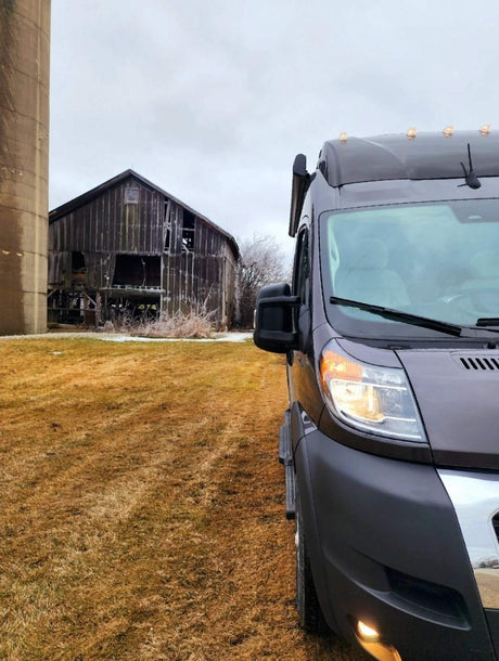 Grey RV rental van on dry grass, with a rustic, dilapidated barn and silo under an overcast sky.