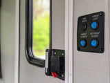 RV rental interior: Light control panel with blue buttons for porch, cabinet, and exterior accent lights, plus a black main light switch, beside a window.