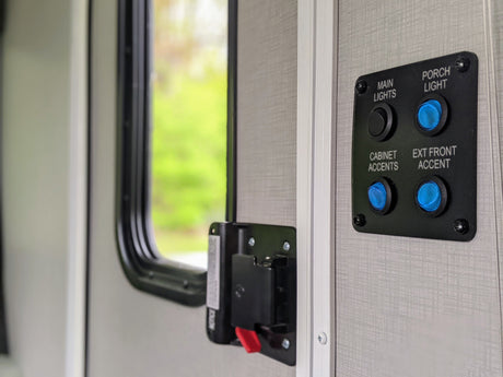 RV rental interior: Light control panel with blue buttons for porch, cabinet, and exterior accent lights, plus a black main light switch, beside a window.