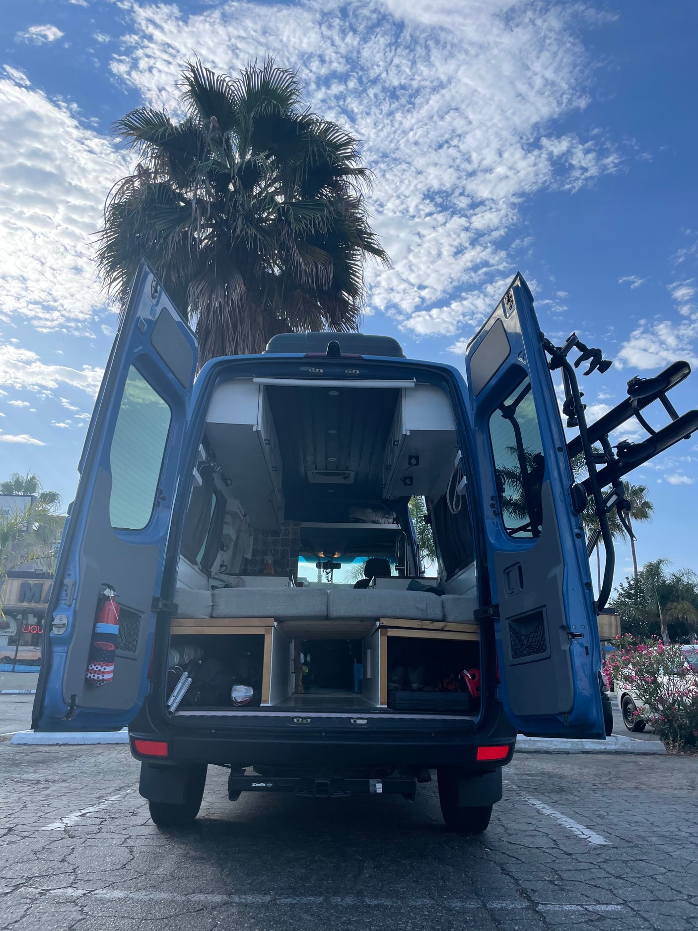 Adventure-ready blue RV rental with open rear doors showing bed, storage, and bike rack under a palm tree. Perfect for your next road trip!