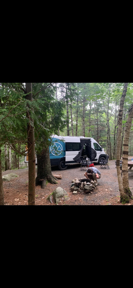 RV rental: Man prepares fire at wooded campsite with a camper van.