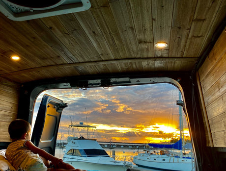 Enjoy a stunning marina sunset from this cozy RV rental! A child views boats from inside the wood-paneled camper van.
