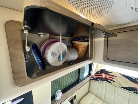 Inside an RV rental: an open overhead cabinet stores kitchen items like paper plates & bowls. A window, fan, and blanket are visible.