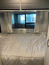 RV rental interior: cozy bed with open curtains, revealing Airstream trailers outside a large window. Overhead blinds are closed.