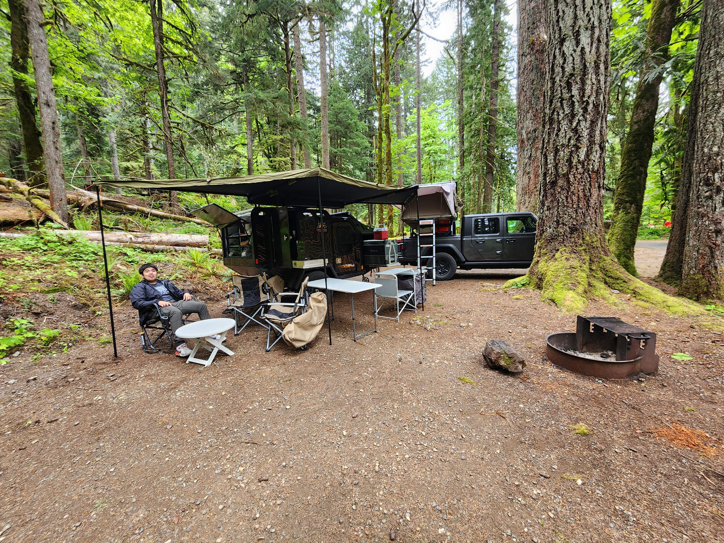 An RV rental overlanding adventure at a forest campsite. A man relaxes beside a rugged trailer & Jeep with a rooftop tent.
