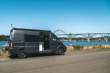 Explore with an **rv rental**! Gray camper van with open door by blue water, featuring a large scenic bridge backdrop.