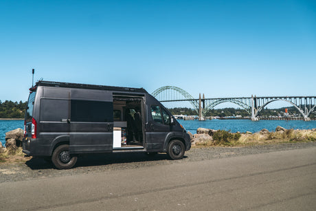 Explore with an **rv rental**! Gray camper van with open door by blue water, featuring a large scenic bridge backdrop.