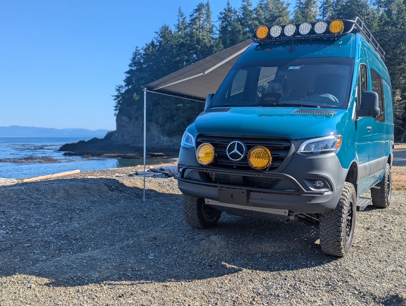 A teal Mercedes-Benz Sprinter camper van, an ideal rv rental, is parked on a rocky coast with an awning extended, facing the ocean.