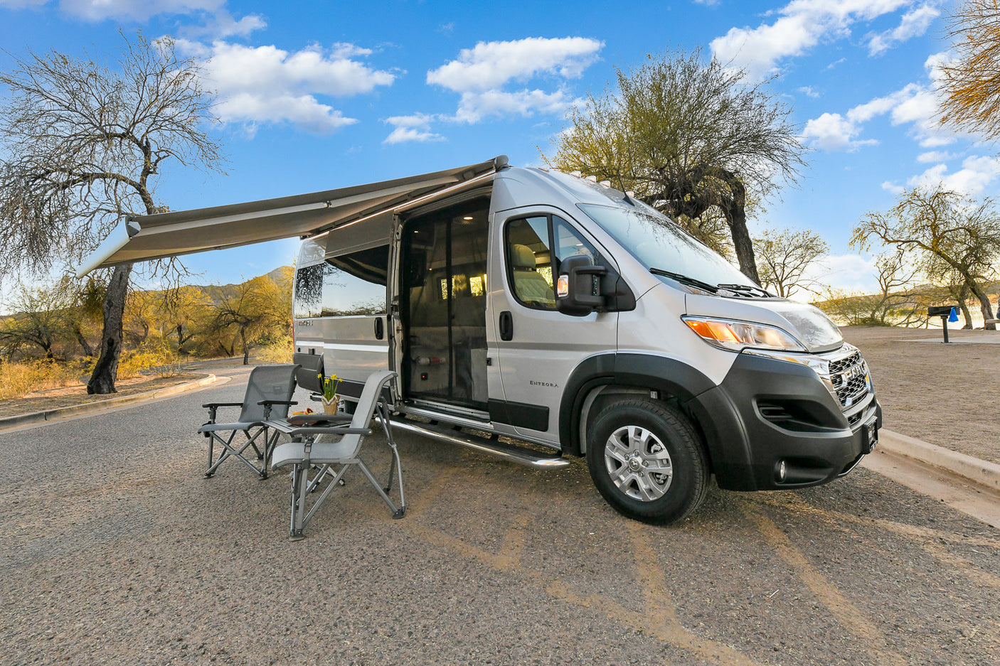 Silver camper van RV rental with awning extended and chairs set for outdoor leisure.