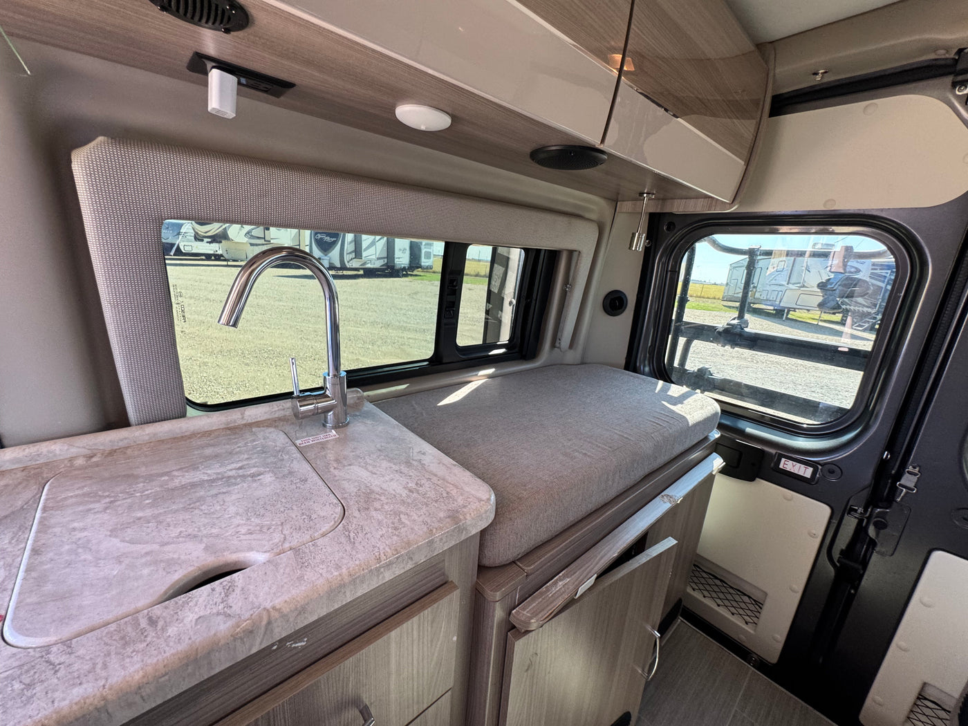 RV rental interior: A compact kitchenette with a sink, countertop, and an adjacent cushioned bench by the window, overlooking other RVs. — photo 8