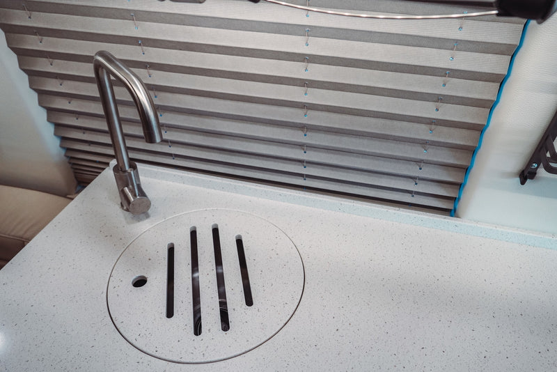 Modern RV rental interior featuring a white speckled countertop with a sleek stainless faucet and a unique slotted drain, next to pleated blinds. — photo 18