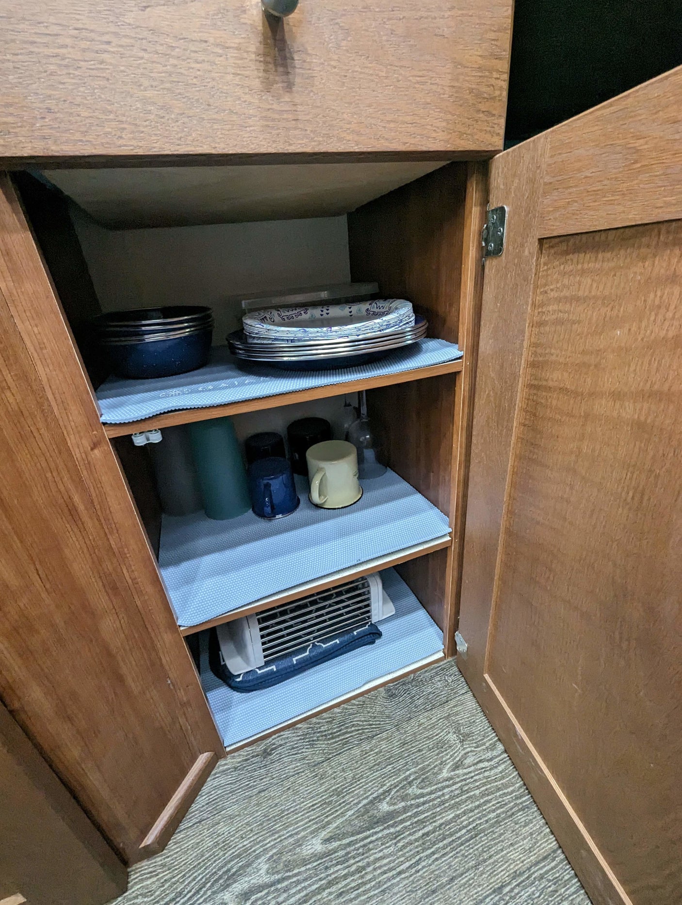 Inside an RV rental kitchen cabinet: blue shelf liners, neatly stacked dishes, mugs, and a compact appliance.