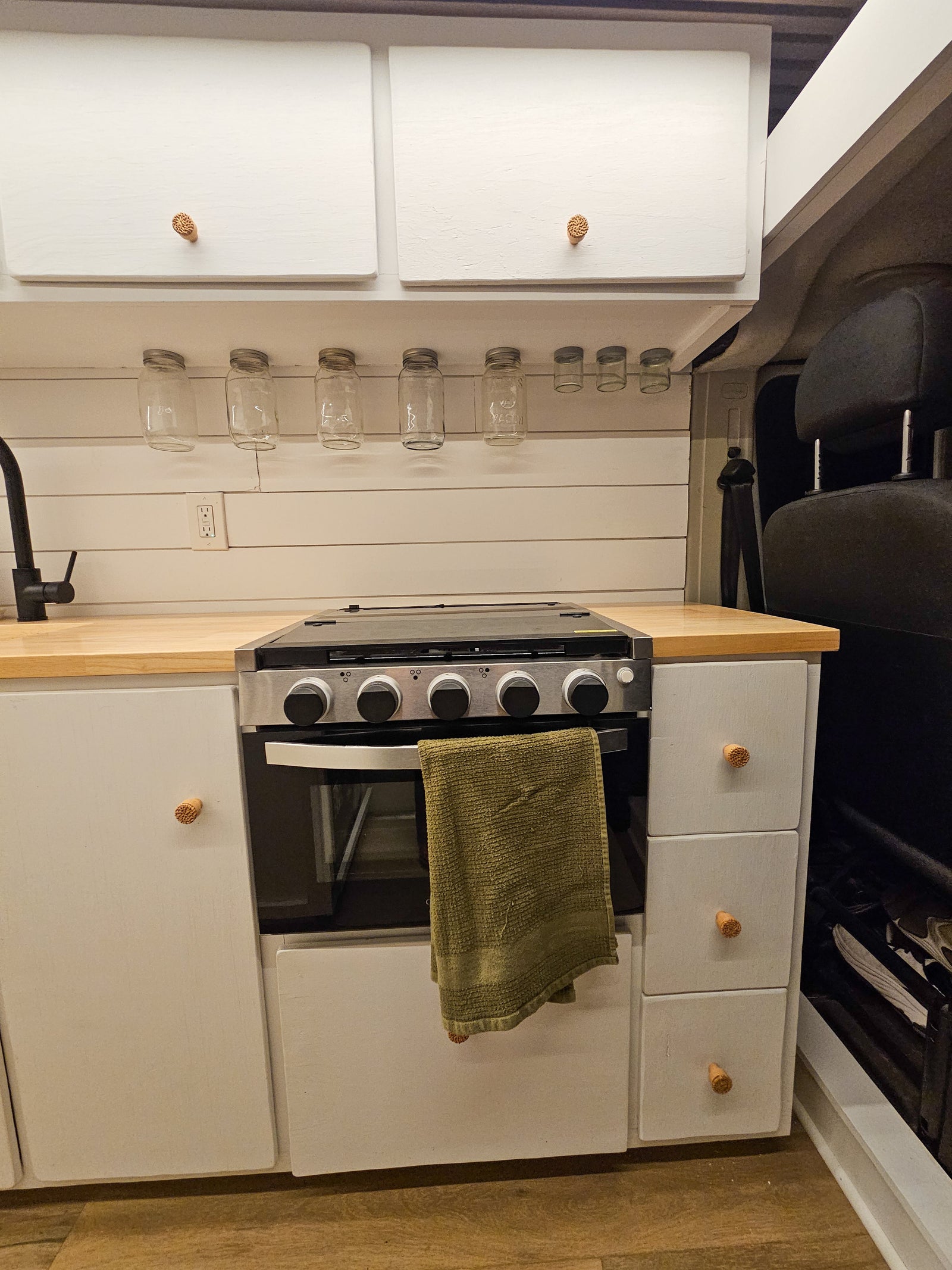 Compact RV rental kitchen features white cabinets, wood counter, a stove, and hanging spice jars.