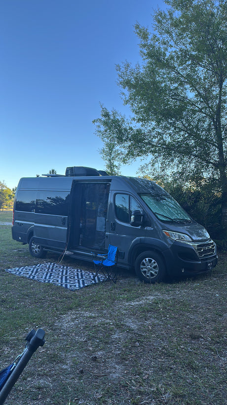 A dark gray camper van, an rv rental, parked for camping with its side door open, a patterned mat, and blue chair.