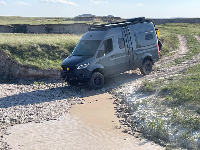 Rugged grey RV rental van drives through a muddy creek in a grassy — photo 1