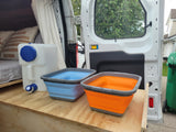 RV rental camper van interior with a bed, water tank, and colorful collapsible bins on a wooden platform.